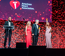Photo of four people standing on stage below a large projection of the American Heart Association heart and torch logo.